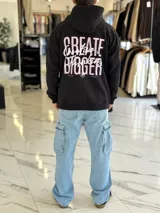 Canguro oversize de color beige claro con capucha, mangas largas y puños elastizados. Presenta un diseño gráfico en la espalda con el texto 'Creative Thinking' en tipografía cursiva negra sobre un fondo de letras grandes en tono rosado.