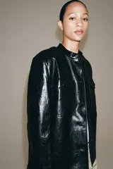 Cazadora efecto piel de Zara Woman Collection, en color negro. Cuenta con cuello redondo, manga larga, bolsillos de parche con solapa en el delantero y cierre frontal con botones forrados. Incluye un cinturón del mismo tejido para ajustar en la cintura.