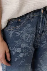 Jean de tiro medio con calce wide leg y pierna amplia, confeccionado en denim azul con estampado floral tono sobre tono. Cuenta con bolsillos delanteros y traseros, y cierre frontal con botón y cremallera.