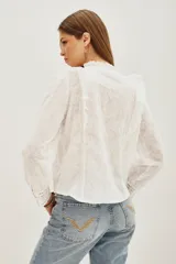 Camisa blanca de algodón con diseño romántico, cuello alto con volados, cierre frontal de botones y detalles de encaje y bordados calados. Presenta mangas largas con puños de encaje y volados frontales.