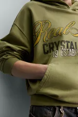 Sudadera con capucha de corte relajado en color verde oliva con efecto lavado. Presenta un bolsillo frontal tipo canguro y un estampado gráfico en el pecho con letras en tonos amarillos y negros. Cuenta con puños y bajo acanalados.