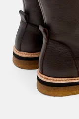 Bota de caña media confeccionada en cuero vacuno texturizado color marrón oscuro. Presenta un diseño minimalista con costuras reforzadas, tirador trasero y una suela de plataforma plana con capas en tonos beige, negro y suela.