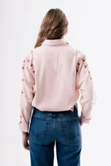 Camisa de algodón color rosado con cuello clásico y cierre frontal de botones. Presenta detalles de bordados florales en tonos rojizos a lo largo de las mangas y costuras con encaje decorativo en los hombros.