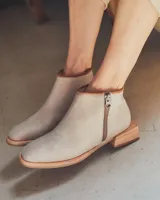 Botinetas de cuero en tono gris claro, con diseño de caña baja, cierre lateral metálico y taco de madera con forma geométrica.