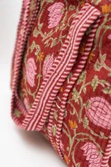 Bolso tipo tote acolchado con estampado floral en tonos rojizos, rosados y verdes. Presenta asas de mano con diseño de rayas verticales en color rojo y blanco, y ribetes a juego en los bordes.
