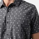 Camisa de manga corta color gris oscuro con estampado de palmeras blancas. Tiene cuello clásico, cierre con botones y bolsillo en el pecho.