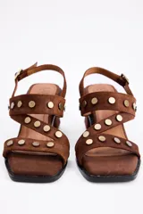 Sandalia de taco cuadrado medio, confeccionada en cuero vacuno negro. Presenta tiras cruzadas sobre el empeine decoradas con tachas metálicas doradas y cierre con hebilla ajustable en el tobillo.