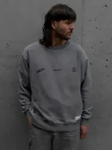 Buzo oversize de cuello redondo en color gris, con diseño estampado en el frente que incluye logotipos y texto. Presenta puños y ruedo acanalados, con una pequeña etiqueta decorativa en la parte inferior.