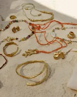 Collar corto de estilo artesanal, compuesto por una variedad de piedras naturales en tonos tierra, verdes, rojos y amarillos, intercaladas con pequeñas perlas de río. Presenta dijes de bronce macizo con formas orgánicas, incluyendo una figura de pez y un dije circular central, unidos por un cierre de barra y argolla.