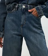 Pantalón de jean modelo carrot con cintura alta, corte holgado en caderas y piernas que se estrechan hacia el tobillo. Confeccionado en denim de algodón con lavado medio, cuenta con cinco bolsillos funcionales, cierre frontal con botón y cremallera, y trabillas para cinturón.