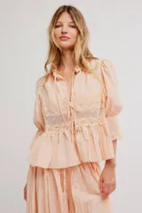 Camiseta de tela liviana color salmón, con terminaciones de encaje, escote en V profundo y botonadura al frente con tiras para anudar. Mangas tres cuartos abullonadas con puño elastizado y ruedo acampanado, cuello con volado.