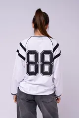 Buzo estilo jersey universitario de color blanco con detalles en negro. Presenta cuello en V, mangas largas con franjas negras y un número estampado en la espalda. Confeccionado en tela deportiva, con corte holgado y puños acanalados.