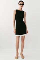 Vestido corto de punto canalé negro, con cuello barco, sin mangas y detalle de volados blancos en el borde inferior.