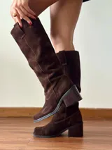 Botas de caña alta confeccionadas en cuero gamuzado color chocolate. Presentan un diseño de estilo western con puntera cuadrada, taco bloque de altura media y un estribo desmontable con argolla metálica en la zona del tobillo.