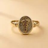 Anillo tipo sello con diseño ovalado que presenta el símbolo del caduceo de la medicina en relieve. La pieza combina plata 925 con detalles en oro 18k, destacando por su acabado texturizado en el fondo del sello.