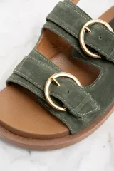 Sandalias de gamuza color verde militar con dos tiras anchas sobre el empeine y una tira trasera, todas ajustables con hebillas doradas. Suela plana color marrón.