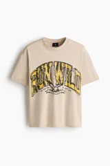 Remera de algodón en color beige con cuello redondo acanalado, hombros caídos y corte holgado. Presenta un estampado frontal con la frase 'RUN WILD' en tonos amarillos y negros con efecto desgastado.
