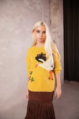 Sweater de punto color amarillo, de calce holgado con cuello redondo y mangas largas. Presenta un diseño bordado a mano en el frente que representa una cigüeña y detalles vegetales.