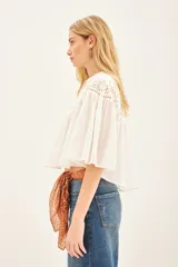 Camisa blanca de corte amplio y diseño cropped, confeccionada en algodón con detalles de encaje bordado en la parte superior y mangas acampanadas con textura rayada.