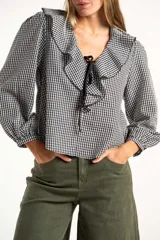 Blusa de manga larga con estampado de cuadros vichy en blanco y negro. Presenta un cuello con volados amplios y un detalle de lazo negro en el centro del escote.