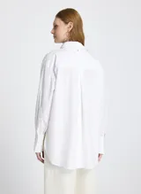 Camisa blanca de corte recto y estilo oversized, con cuello clásico, mangas largas con puños anchos, bolsillo delantero, cierre frontal con botones ocultos y bajo ligeramente redondeado.