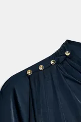 Blusa de tejido satinado color bordó, con cuello subido drapeado y manga larga con puños. Presenta detalle de pliegues frontales y cierre con botones laterales en el cuello.