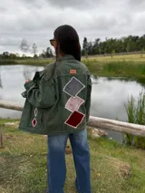 Campera de jean color verde oliva, con diseño de figuras geométricas en terciopelo aplicadas en la espalda y mangas, bordadas a mano con costuras decorativas. Presenta un corte holgado y etiqueta de cuero en la parte superior de la espalda.