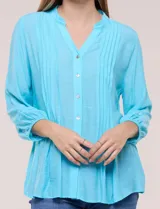 Blusa celeste de silueta suelta y textura liviana, con escote en V, cuello mao y cierre frontal de botones. Cuenta con alforzas verticales en el pecho y mangas tres cuartos con puños elastizados y detalle de galón calado.