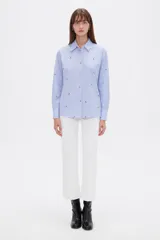 Blusa de corte holgado con cuello camisero y cierre frontal de botones. Presenta un diseño de rayas verticales finas en color azul y blanco, decorado con pequeños bordados dispersos.