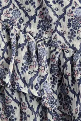 Vestido corto de tejido transparente, con estampado de patrón geométrico y floral en tonos azules y blancos, cuello redondo con pequeña abertura y tiras para atar. Tiene manga larga abullonada, cintura elastizada y falda con volados en capas.