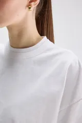 Remera cropped blanca de calce relajado, con cuello redondo, hombros caídos y mangas cortas. Presenta un pequeño detalle de etiqueta bordada en el lateral inferior.