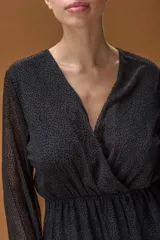 Vestido corto negro de tela semitransparente con estampado de micro lunares. Presenta escote en V cruzado, cintura elástica, mangas largas con puños ajustados y falda con volados en capas.