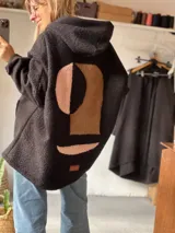 Campera tipo hoodie de textura tipo corderito en color negro, con diseño de collage abstracto en la espalda compuesto por formas geométricas en tonos marrón y rosado. Presenta capucha, mangas largas y un corte holgado.