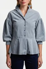 Camisa de poliéster color gris azulado, con mangas abullonadas, cuello mao con volados y corte peplum en la cintura. Presenta cierre frontal con botones.