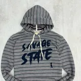 Canguro gris con rayas horizontales en tonos gris, rosa y blanco. Estampado frontal con la frase "Savage State" en letras negras. Tiene capucha con cordones blancos, bolsillo tipo canguro y puños negros.