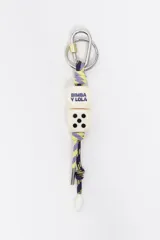Llavero de cordón trenzado en tonos violeta y amarillo, con argolla metálica y mosquetón plateado. Presenta dos cuentas en forma de dado de color crudo, una con la inscripción de la marca en azul y la otra con puntos negros.