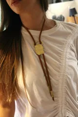 Corbatín con cordón de cuero marrón y dije de bronce dorado con forma irregular.