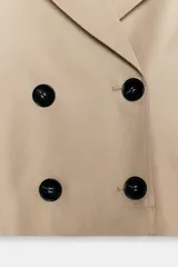 Trench corto de color beige con cuello de solapa, cierre frontal cruzado con botones y mangas largas con trabillas ajustables en los puños. Presenta un diseño estructurado con hombreras y bolsillos delanteros de vivo.