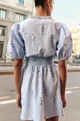 Vestido corto de color celeste con estampado floral, cuello redondo con volante y escote en pico. Presenta mangas cortas abullonadas con puños elásticos y cintura con nido de abeja elástico.
