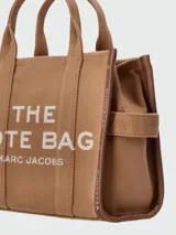 Bolso tipo tote de lona color negro con letras blancas bordadas en el frente que dicen 'THE TOTE BAG MARC JACOBS'. Cuenta con asas superiores cortas y una correa de hombro ajustable y extraíble.