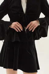 Saco de terciopelo negro con cierre frontal mediante lazos, cuello mao, mangas largas con puños acampanados y detalle de volado en la cintura.