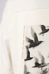 Remera de corte oversize color crudo, con cuello redondo y mangas cortas. Presenta un pequeño estampado gráfico en el pecho con diseño de aves en vuelo.