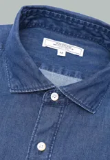 Camisa de jean azul con botones blancos, mangas largas con puños doblados y corte clásico.