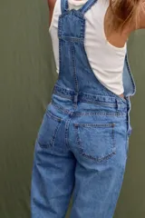 Jumpsuit de jean azul clásico con diseño de jardinera, tirantes ajustables con hebillas metálicas, bolsillo frontal con etiqueta de marca y bolsillos laterales.