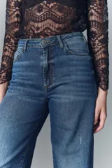 Jean de corte oxford confeccionado en denim azul con lavado desgastado y bigotes. Presenta tiro alto, pierna ancha que se ensancha desde la cadera hacia el ruedo, cierre frontal con botón y pequeñas roturas decorativas.