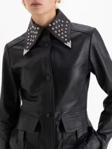 Parka de cuero vacuno color negro, con diseño utilitario que incluye bolsillos frontales con tapa y fuelle. Presenta un cuello con detalle de tachas metálicas y cierres en los puños.