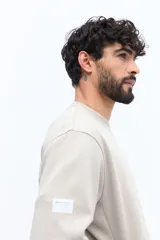 Buzo de algodón y poliéster color verde musgo, con cuello redondo y puños acanalados. Presenta un parche estampado rectangular con texto y un logo circular en el pecho.