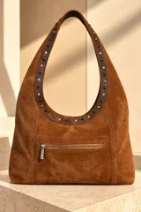 Cartera tipo hobo de gamuza color marrón chocolate, con tira de cuero decorada con tachas metálicas doradas y negras. Incluye placa metálica con el logo de la marca en la base.