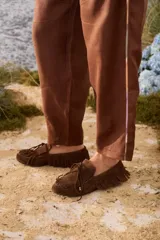 Mocasines de gamuza color tostado, con diseño clásico, flecos alrededor del calzado y en la capellada, y cordón decorativo con detalles metálicos dorados.