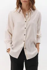 Camisa beige de manga larga con botones marrones y cuello clásico. Presenta un detalle de costura dorada a lo largo de la botonera.